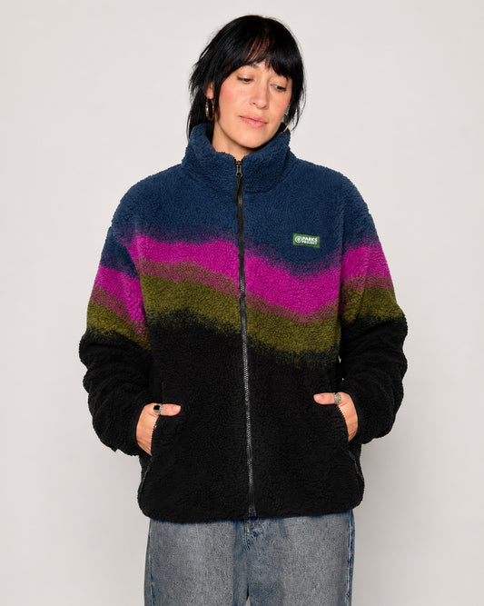 A woman wearing a fuzzy quarter-zip fleece jacket with horizontal wavy stripes in navy blue, magenta, olive green, and black, paired with light-wash jeans against a plain light gray background.
Shop Borealis High Pile Fleece Jacket Inspired by Alaskan Parks | multi-color