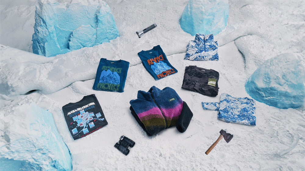 Image of 8 different items inspired by Alaska as the Last Frontier. Items include Borealis inspired art, mountain scape prints, and printed art of the 8 Alaskan National Parks. All folded and laying on a snowy background with ice chunks