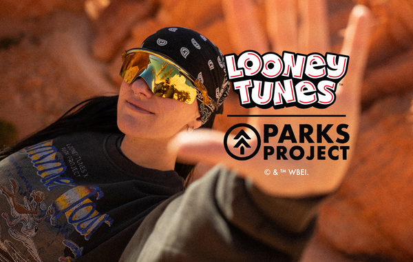Person reclining among warm-toned desert rock formations, wearing reflective sunglasses, a black bandana, and a graphic sweatshirt, with one hand raised toward the camera in a sunlit outdoor setting. Looney Tunes and Parks Project co-branded logo displayed on a dark background, featuring the Looney Tunes wordmark below the Parks Project logo and brand name. © & ™ WBEI.