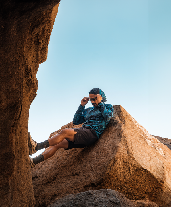 Person wearing a blue hooded sun shirt and sunglasses relaxing on large desert rock formations under a clear blue sky.