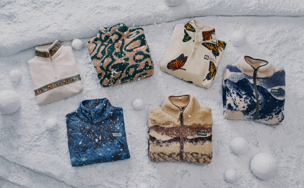 An image of six fleece jackets arranged on a snowy surface with snowballs scattered around. The jackets feature various patterns, including a cream-colored jacket with a patterned collar, a green and beige animal print design, a white jacket with butterfly prints, a blue jacket with a mountain-like design, and a beige and brown jacket with a wave-like design.