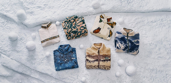 An image of six fleece jackets arranged on a snowy surface with snowballs scattered around. The jackets feature various patterns, including a cream-colored jacket with a patterned collar, a green and beige animal print design, a white jacket with butterfly prints, a blue jacket with a mountain-like design, and a beige and brown jacket with a wave-like design.