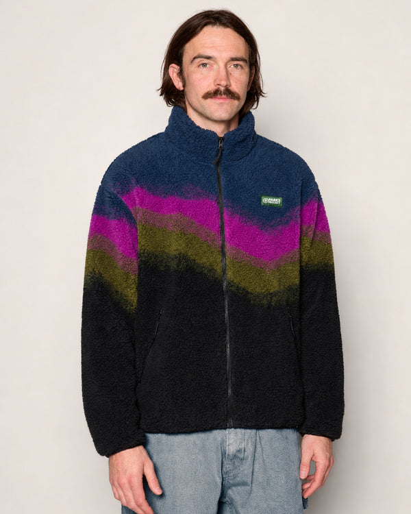 A man wearing a Parks Project fleece jacket with a dark navy base and wavy stripes of magenta, olive green, and black, paired with light blue jeans against a plain beige background.

Shop Borealis High Pile Fleece Jacket Inspired by Alaskan Parks | multi-color