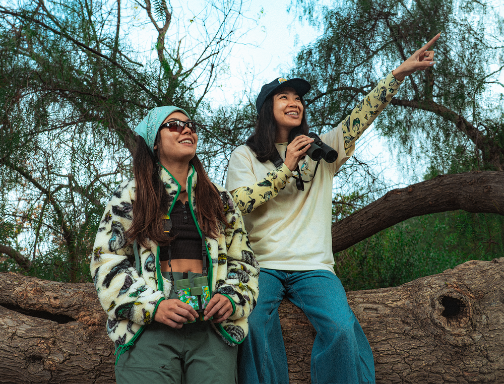 Two outdoor enthusiasts sit on a large fallen tree beneath leafy branches. One person holds binoculars and points toward the sky while the other wears sunglasses and apparel with birds in the all over print and holds a field guide, both engaged in birdwatching. SHOP THE AUDUBON X PARKS PROJECT COLLECTION