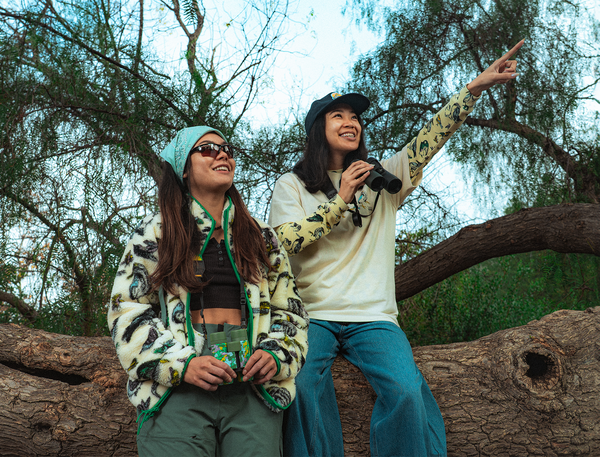Two outdoor enthusiasts sit on a large fallen tree beneath leafy branches. One person holds binoculars and points toward the sky while the other wears sunglasses and apparel with birds in the all over print and holds a field guide, both engaged in birdwatching. SHOP THE AUDUBON X PARKS PROJECT COLLECTION
